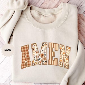 Op de afbeelding: Een zandkleurige sweatshirt met het woord "AMEN" in een lettertype in collegestijl. De letters hebben verschillende patronen, waaronder ruitjes, bloemen, strepen en luipaardprint. Een bijpassende scrunchie is over de schouder gedrapeerd.