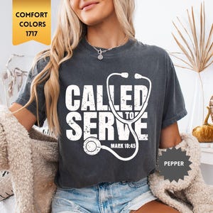 Può includere: T-shirt grigio scuro con la scritta "CALLED TO SERVE" e una grafica di stetoscopio bianca. Il testo "MARK 10:45" è in basso. La maglietta è in stile Comfort Colors 1717.