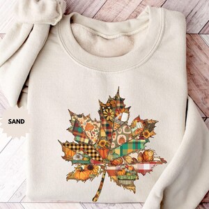 May include: A sand-coloured sweatshirt featuring a large, colourful leaf design. The leaf is a patchwork of autumn-themed patterns, including pumpkins, sunflowers, and plaid. The word "SAND" is printed on the left side of the sweatshirt.