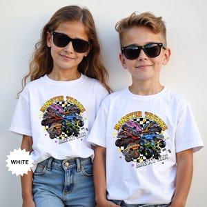 May include: Two children wearing white t-shirts with a colorful monster truck graphic. The graphic features various monster trucks arranged around a checkered circle with the text "Monster Trucks Are My Jam". The shirts are paired with blue jeans.