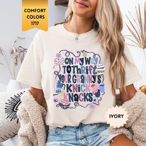 Może przedstawiać: Kremowa koszulka z napisem "ON MY WAY TO THRIFT YOUR GRANNY'S KNICK KNACKS" w kolorowym, fantazyjnym wzorze. Koszulka jest w stylu Comfort Colors 1717.