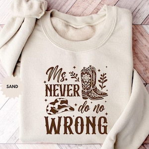 Puede incluir: Una sudadera de color arena con el texto "Ms. Never do no Wrong" en marrón. El diseño incluye un sombrero de vaquero, botas de vaquero y detalles florales. La sudadera tiene cuello redondo y mangas largas.