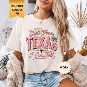 May include: Ivory-colored t-shirt with the text "She's From Texas I Can Tell" in pink and turquoise. The shirt has a relaxed fit and short sleeves. The words "Comfort Colors" and "Ivory" are also visible.