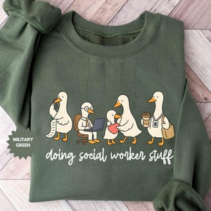 Könnte beinhalten: Militärgrünes Sweatshirt mit einer Grafik von Cartoon-Enten, die jeweils mit Aufgaben eines Sozialarbeiters beschäftigt sind. Der Schriftzug "doing social worker stuff" ist in weißer Schrift geschrieben. Das Sweatshirt hat einen Rundhalsausschnitt und lange Ärmel.