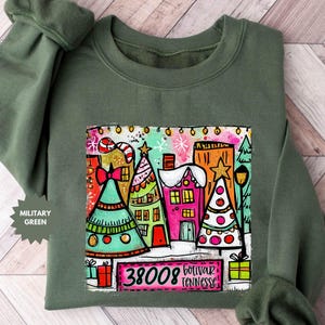 May include: Military green sweatshirt with a colorful Christmas-themed graphic. The graphic features stylized houses, Christmas trees, and the text "38008 Bolivar Tennessee". The sweatshirt has a crew neck and long sleeves.