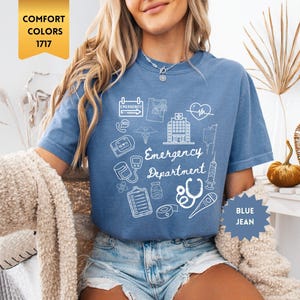May include: Blue jean t-shirt with white graphic illustrations of medical symbols and the words "Emergency Department". The shirt has short sleeves and a crew neck. The shirt is a comfort color 1717.