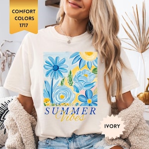 Peut inclure: Un t-shirt blanc avec un motif de fleurs bleues et jaunes et le texte "Summer Vibes". Le t-shirt est port&eacute; par un mannequin portant un short en jean bleu. Le t-shirt est &eacute;tiquet&eacute; "Ivoire".