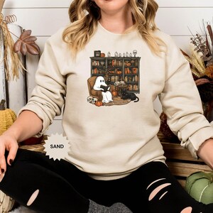 May include: A sand-colored sweatshirt with a graphic of a ghost reading a book in a chair next to a bookshelf filled with books. The ghost is wearing a black hat and the chair is next to a black cat. The text "SAND" is printed on the sweatshirt.