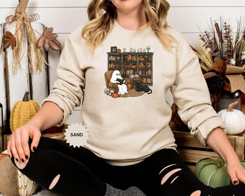 May include: A sand-colored sweatshirt with a graphic of a ghost reading a book in a chair next to a bookshelf filled with books. The ghost is wearing a black hat and the chair is next to a black cat. The text "SAND" is printed on the sweatshirt.
