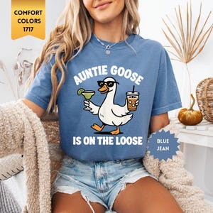 May include: A blue jean coloured t-shirt featuring a graphic of a white goose wearing black sunglasses. The goose holds a margarita in one wing and an iced coffee in the other. The text reads AUNTIE GOOSE IS ON THE LOOSE in white lettering.