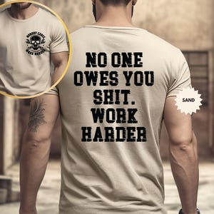 Op de afbeelding: Zandkleurig T-shirt met de zwarte tekst "NO ONE OWES YOU SHIT. WORK HARDER" op de achterkant. Een kleinere afbeelding op de voorkant toont een schedel met sleutels en de woorden "NOBODY CARES WORK HARDER".
