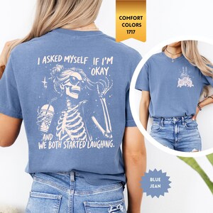 May include: Blue jean-coloured t-shirt featuring a skeleton graphic. The skeleton wears sunglasses and holds a drink. The text on the shirt says, "I asked myself if I'm okay, and we both started laughing."