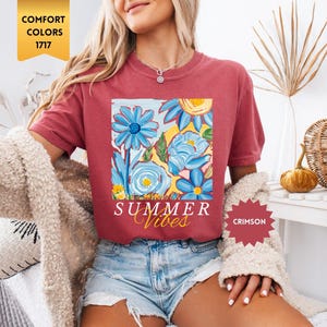 Peut inclure: Un t-shirt rouge avec un motif floral et le texte "Summer Vibes" en blanc. Le t-shirt est port&eacute; par une personne aux cheveux blonds. Le texte "Crimson" est visible dans un cercle rouge.