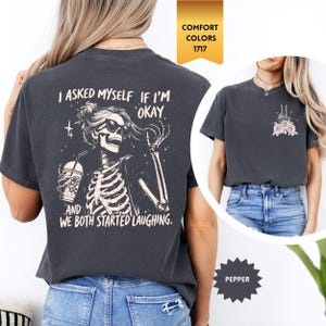 May include: Dark grey t-shirt with a skeleton graphic wearing sunglasses and holding a drink. The text on the back reads "I asked myself if I'm okay, and we both started laughing." The shirt is a Comfort Colors 1717 in Pepper.