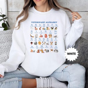 May include: White sweatshirt featuring the text "VETERINARY ALPHABET" with an illustrated alphabet of animals. The design includes various animals, such as dogs, cats, and birds, each representing a letter of the alphabet. A white starburst graphic with the word "WHITE" is also visible.