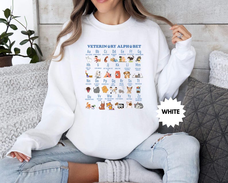May include: White sweatshirt featuring the text "VETERINARY ALPHABET" with an illustrated alphabet of animals. The design includes various animals, such as dogs, cats, and birds, each representing a letter of the alphabet. A white starburst graphic with the word "WHITE" is also visible.