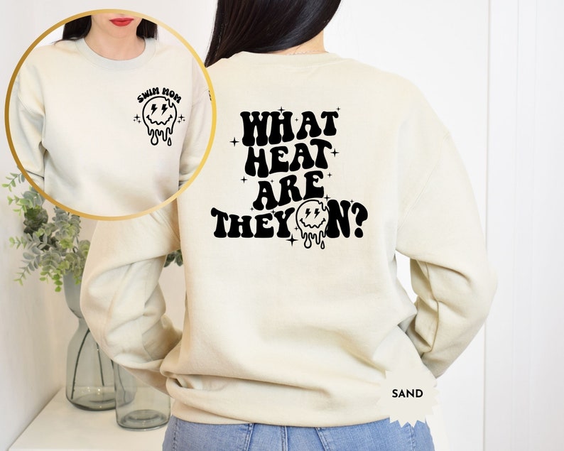 May include: A cream coloured sweatshirt with a black graphic on the back that says "WHAT HEAT ARE THEY ON?" with a dripping smiley face. The front of the sweatshirt has a black graphic of a dripping smiley face with the text "SWIM MOM".