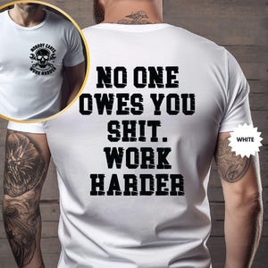 Op de afbeelding: Wit T-shirt met de tekst "NO ONE OWES YOU SHIT. WORK HARDER" in vette zwarte letters op de achterkant. De voorkant van het shirt heeft een schedel- en moersleutelafbeelding met de woorden "NOBODY CARES WORK HARDER".