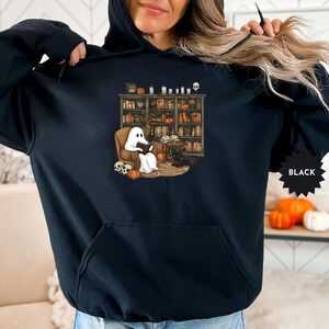 May include: Black hoodie with a graphic of a ghost reading a book in a library. The ghost is sitting in a chair with a book in its hands. The library has bookshelves filled with books and a skull on the floor.