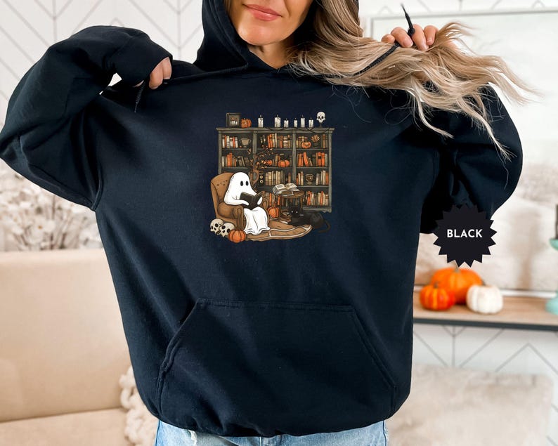 May include: Black hoodie with a graphic of a ghost reading a book in a library. The ghost is sitting in a chair with a book in its hands. The library has bookshelves filled with books and a skull on the floor.