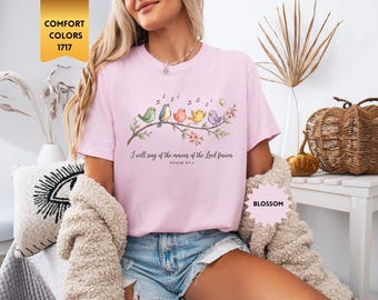 May include: A light pink t-shirt featuring a graphic of five colorful birds perched on a flowering branch with musical notes and a butterfly. The text reads, I will sing of the mercies of the Lord forever, PSALM 89:1. A gold tag says COMFORT COLORS 1717.