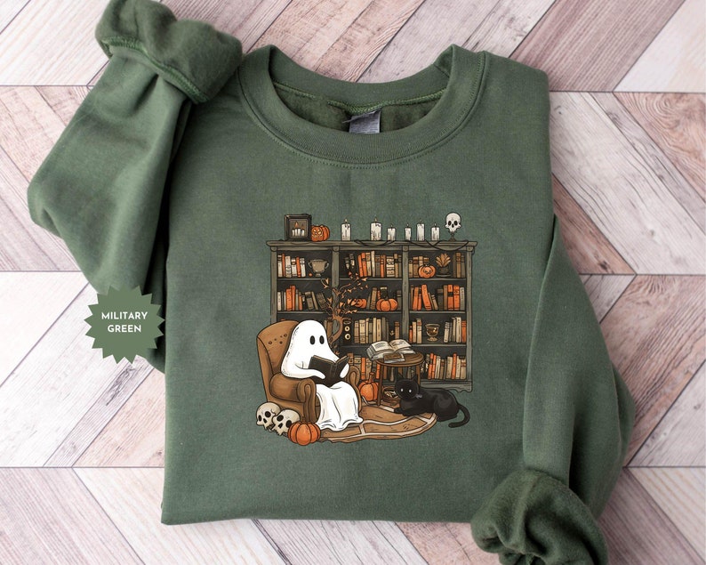May include: A military green sweatshirt with a graphic of a ghost reading a book in a chair in front of a bookshelf with pumpkins and candles. The ghost is wearing a white sheet and has a black book in its hands. The bookshelf is filled with books and has a skull on top.