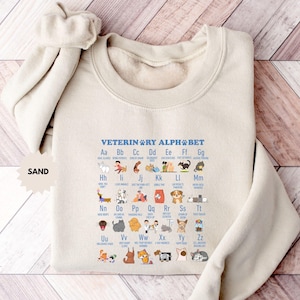 May include: Sand-colored sweatshirt featuring a veterinary alphabet design. The design includes the text "VETERINARY ALPHABET" and illustrations of various animals and veterinary tools. The sweatshirt has a crew neck and long sleeves.