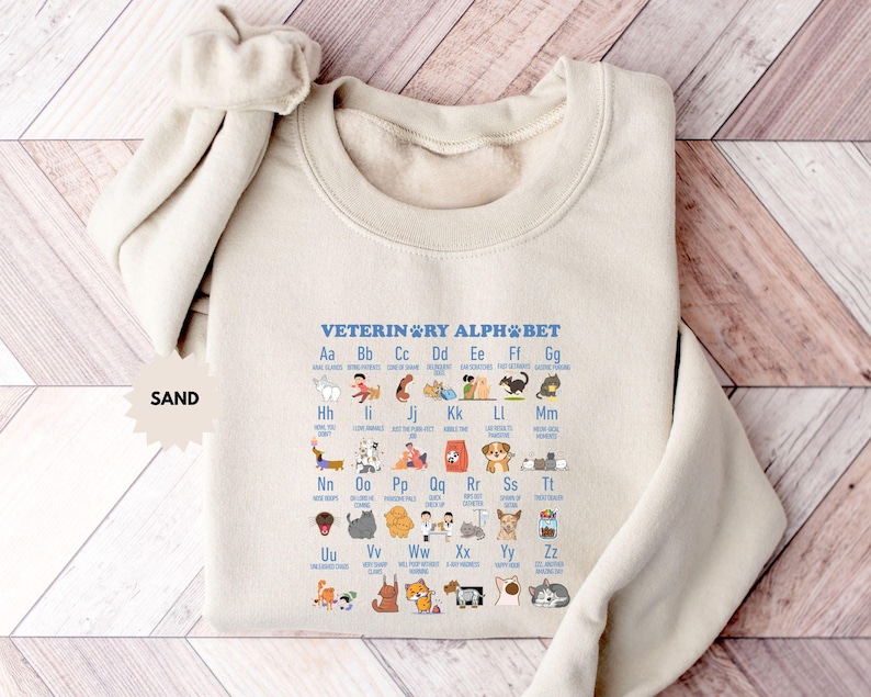May include: Sand-colored sweatshirt featuring a veterinary alphabet design. The design includes the text "VETERINARY ALPHABET" and illustrations of various animals and veterinary tools. The sweatshirt has a crew neck and long sleeves.