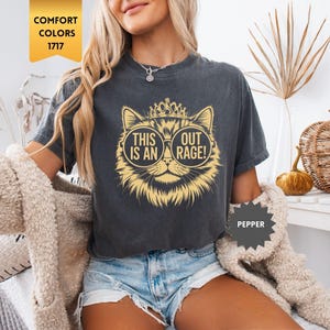 May include: Dark gray t-shirt featuring a cat wearing sunglasses and a crown. The cat graphic has the text "THIS IS AN OUT RAGE!". The shirt is a Comfort Colors 1717 style in the color Pepper.