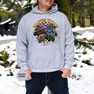 May include: A light gray hooded sweatshirt featuring a colorful graphic of monster trucks. The graphic includes the text "MONSTER TRUCKS ARE MY JAM" in a circular design. The sweatshirt has a kangaroo pocket and a drawstring hood.