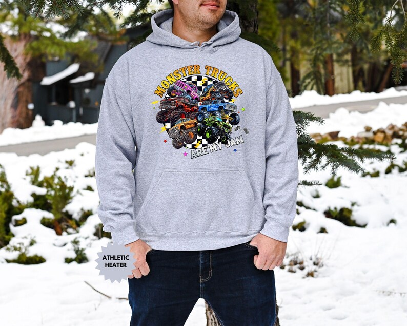 May include: A light gray hooded sweatshirt featuring a colorful graphic of monster trucks. The graphic includes the text "MONSTER TRUCKS ARE MY JAM" in a circular design. The sweatshirt has a kangaroo pocket and a drawstring hood.