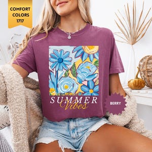 Peut inclure: Un t-shirt violet avec un motif floral et le texte "Summer Vibes" imprim&eacute; sur le devant. Le t-shirt est port&eacute; par une personne portant un short en jean.