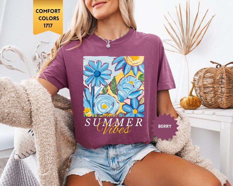 Peut inclure: Un t-shirt violet avec un motif floral et le texte "Summer Vibes" imprim&eacute; sur le devant. Le t-shirt est port&eacute; par une personne portant un short en jean.