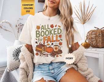 Peut inclure: Un t-shirt ivoire avec le texte ALL BOOKED for FALL en lettres vertes et orange. Le motif comprend une pile de livres attachés par un ruban, une citrouille, une tasse fumante avec une feuille d'érable, une écharpe et des lunettes, entourés de feuilles d'automne.