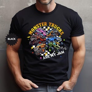 May include: Black t-shirt featuring a colorful graphic of monster trucks. The design includes the text "MONSTER TRUCKS ARE MY JAM" in yellow and white, with a checkered flag and lightning bolt accents. The shirt is black.