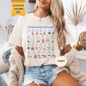 Può includere: Una t-shirt a maniche corte color avorio con una stampa grafica blu dell'alfabeto veterinario. Il design mostra le lettere dalla A alla Z, ciascuna abbinata a una piccola illustrazione di un animale o oggetto veterinario e relativo testo.