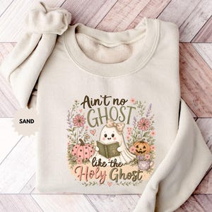 May include: A sand-colored crewneck sweatshirt featuring a graphic of a ghost wearing a bow and reading a book, surrounded by flowers, pumpkins, and the text "Ain't no GHOST like the Holy Ghost" in brown and pink lettering with a small "SAND" label.