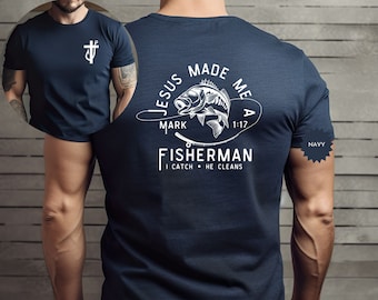 May include: Navy blue t-shirt with a white graphic on the back that reads "JESUS MADE ME A FISHERMAN" with a fish and fishing rod illustration. The front of the shirt has a white cross symbol. The text "I CATCH • HE CLEANS" is also included.