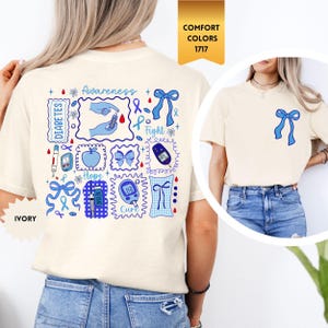 May include: Ivory-colored t-shirt with a blue ribbon graphic on the front and a larger design on the back featuring diabetes awareness symbols, text, and blue ribbons. The shirt has the words "Awareness", "Fight", "Hope", and "Cure".