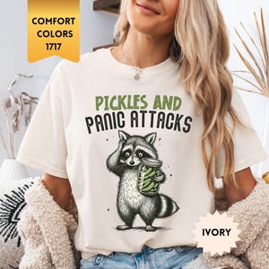 May include: Ivory t-shirt with a raccoon graphic holding a pickle jar. The text "PICKLES AND PANIC ATTACKS" is printed above the raccoon illustration in green and black. The shirt is a comfort color.