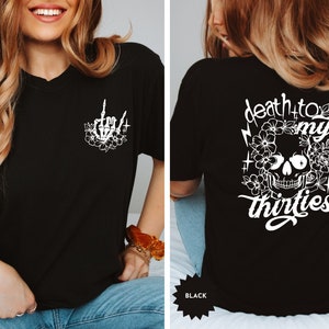 May include: Black t-shirt with a white skull and flowers graphic on the back and a white skeleton hand with flowers graphic on the front. The text on the back reads "death to my thirties".