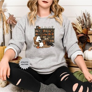 May include: A gray sweatshirt with a ghost reading a book in a library setting. The ghost is white and has a black cat sitting next to it. The text "Athletic Heather" is on the bottom of the sweatshirt.