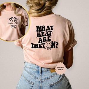 May include: A light pink t-shirt with a black graphic on the front that says "Swim Mom" and a melting smiley face. The back of the shirt has a black graphic that says "What Heat Are They On?" with a melting smiley face.