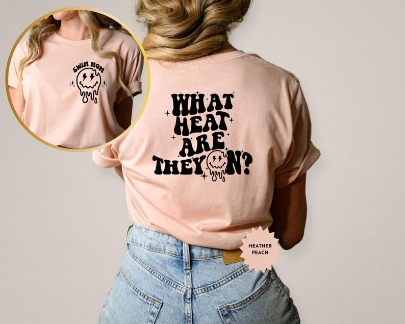 May include: A light pink t-shirt with a black graphic on the front that says "Swim Mom" and a melting smiley face. The back of the shirt has a black graphic that says "What Heat Are They On?" with a melting smiley face.