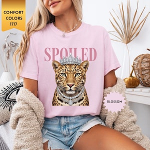 May include: A light pink t-shirt with the word "SPOILED" in pink letters. The shirt features a leopard wearing a crown and a pearl necklace. The shirt is labeled "Blossom" and "Comfort Colors 1717".