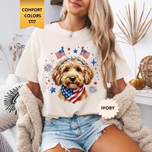 Könnte beinhalten: Elfenbeinfarbenes T-Shirt mit einem patriotischen Hunde-Design. Die Grafik zeigt einen Hund mit einem Bandana und Stirnband mit amerikanischen Flaggen, umgeben von Sternen und Feuerwerk. Das Shirt hat den Text "COMFORT COLORS 1717".