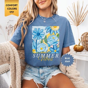 Peut inclure: Un t-shirt bleu avec un motif floral et le texte "Summer Vibes" en jaune. Le t-shirt est port&eacute; par une personne aux cheveux blonds longs et un short en jean bleu.