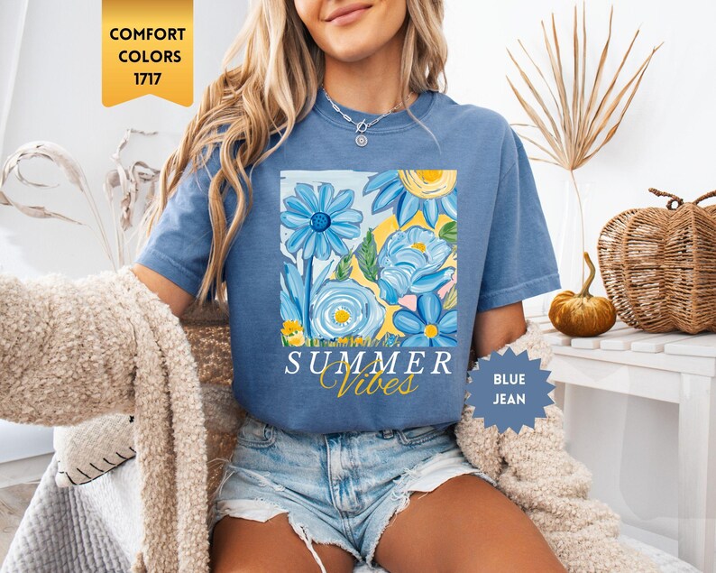 Peut inclure: Un t-shirt bleu avec un motif floral et le texte "Summer Vibes" en jaune. Le t-shirt est port&eacute; par une personne aux cheveux blonds longs et un short en jean bleu.
