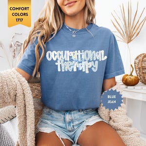 May include: A blue jean colored short-sleeve t-shirt featuring the text "occupational therapy" in a white and light blue floral pattern. The shirt has a crew neckline and a relaxed fit, with a gold tag labeled "COMFORT COLORS 1717" visible.