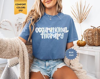 May include: A blue jean colored short-sleeve t-shirt featuring the text "occupational therapy" in a white and light blue floral pattern. The shirt has a crew neckline and a relaxed fit, with a gold tag labeled "COMFORT COLORS 1717" visible.